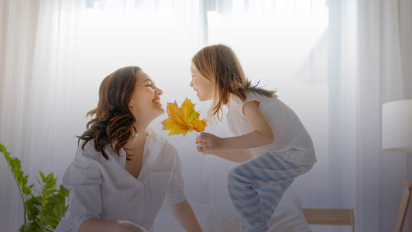image of mother and daughter smiling at each other on a bed with daylight streaming in through a sheer curtain. Daughter is wearing pajamas and holding fall leaves. Used for a blog post about falling asleep naturally with melatonin.
