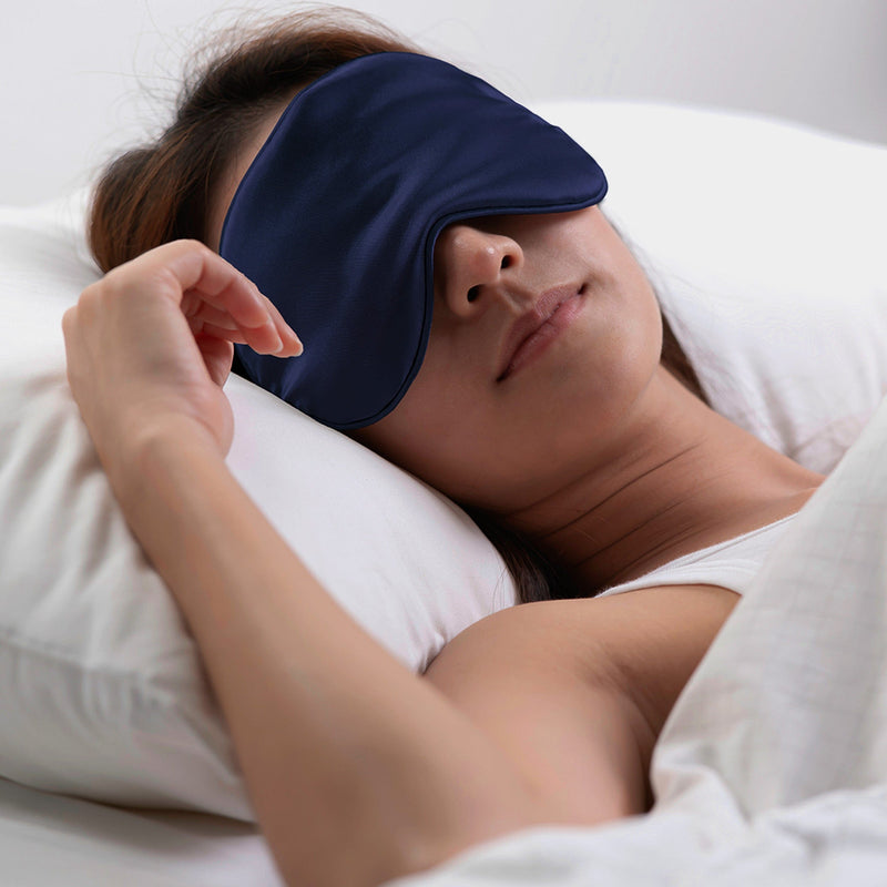 photo of female model in bed, pictured with silk eye mask over her eyes to illustrate how the eye mask is used to block light for better sleep